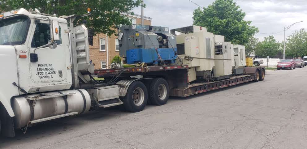 A white truck pulling a lowboy trailer loaded with CNC machines, parked on a street