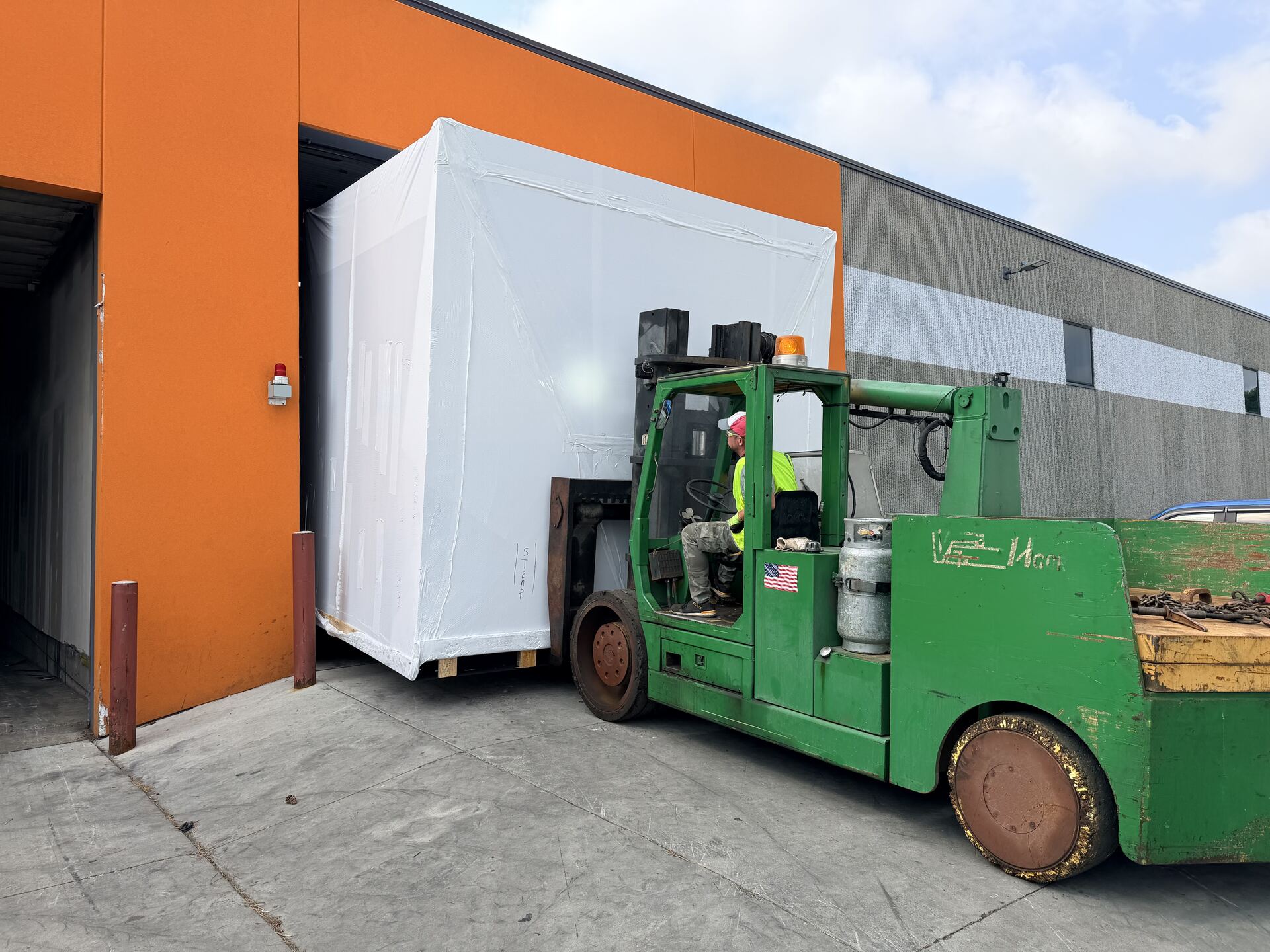 A large green forklift carrying a white-wrapped crate out of a dock door