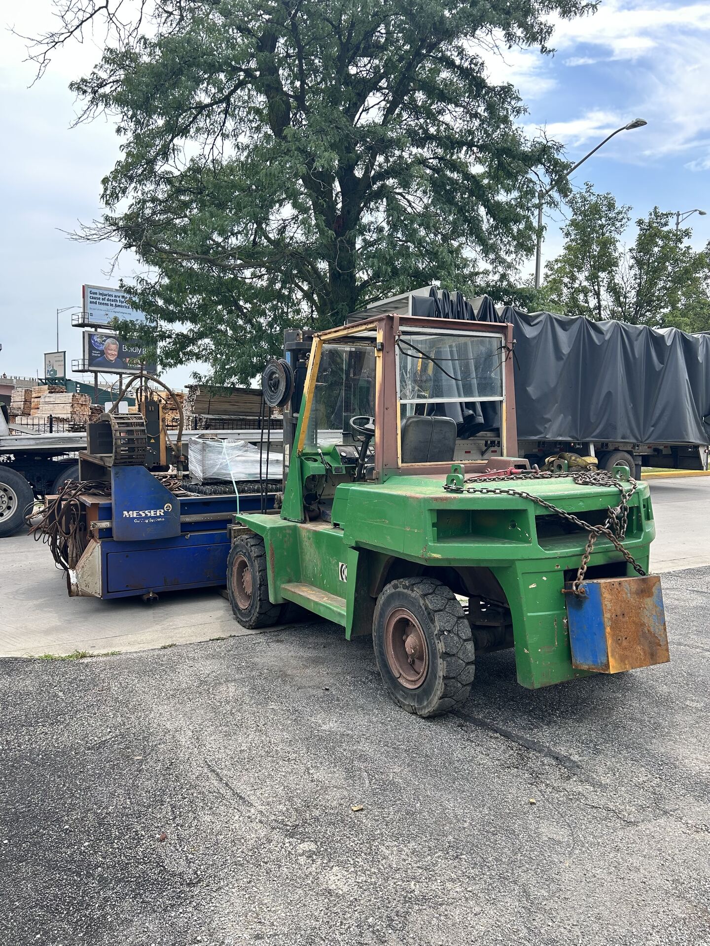 A green forklift parked beside a trailer with a blue machine and a tarped load