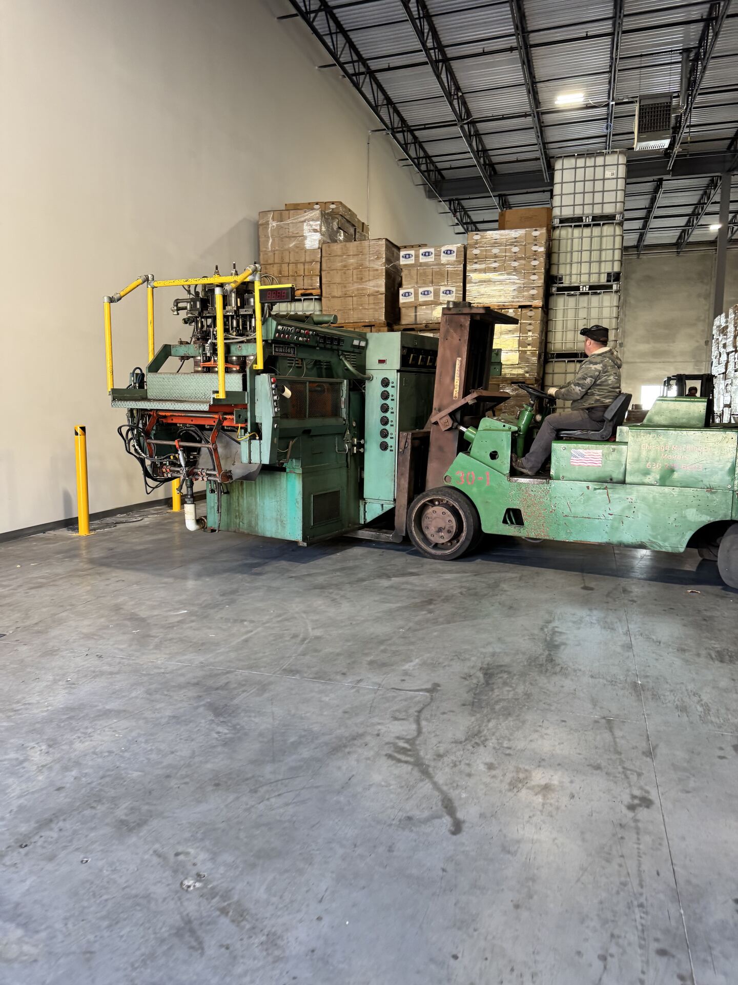 A green forklift carrying a green machine across a warehouse floor