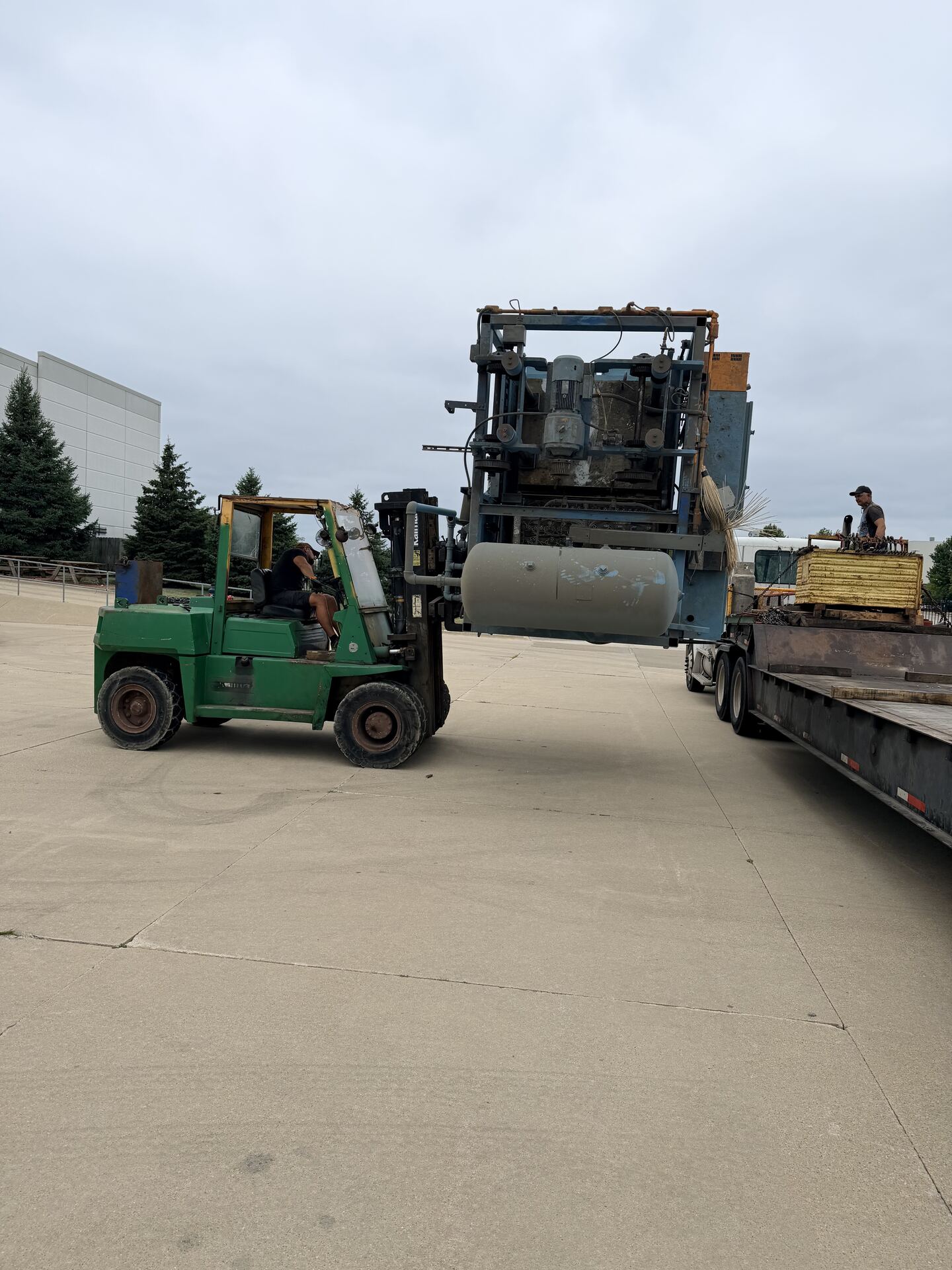 A green forklift lifting a machine beside a flatbed trailer