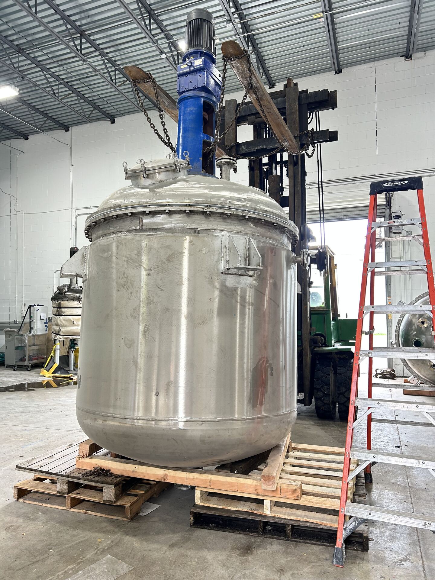 A stainless steel vessel with a blue agitator motor, set on pallets beside a ladder