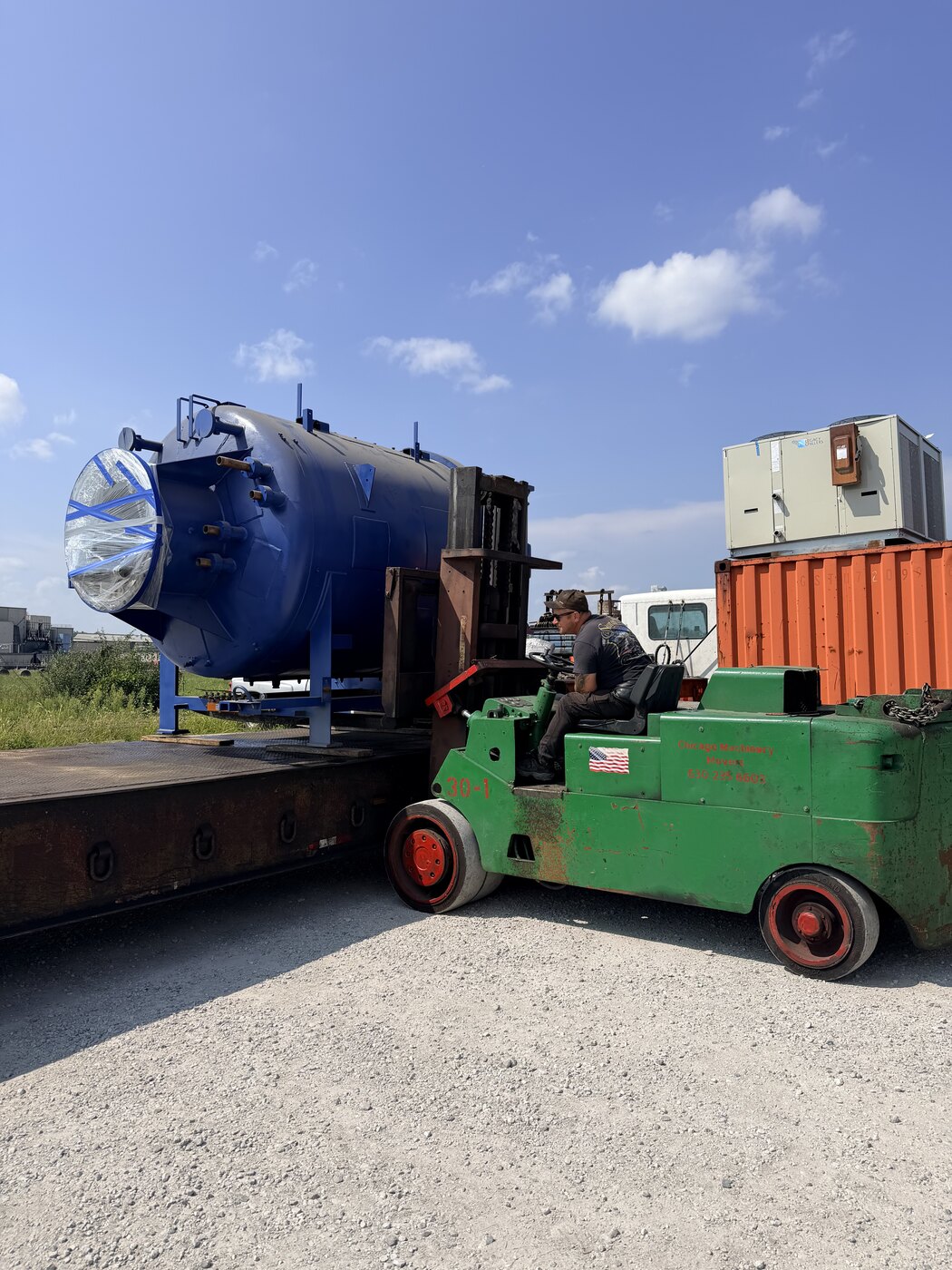 Our green forklift carrying the furnace chamber, the round door end forward, onto the lowboy outside under a blue sky