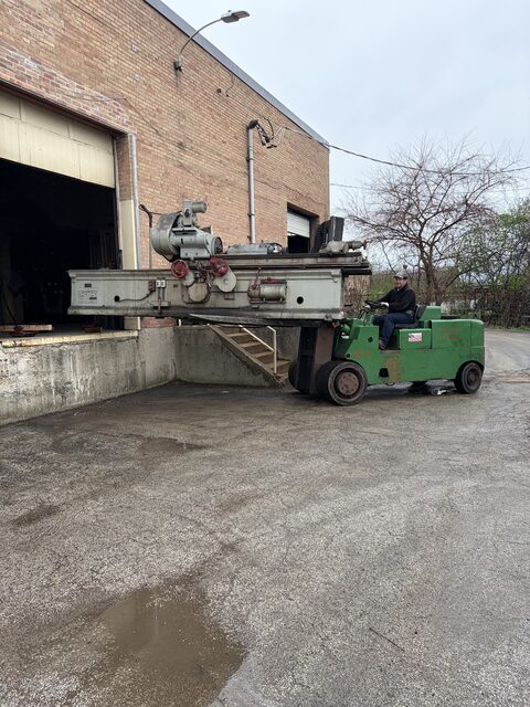 A green forklift carrying a long machine beside a loading dock