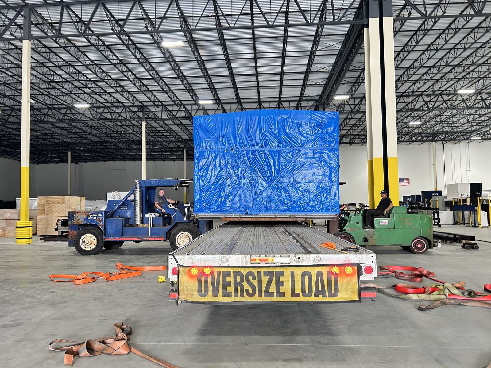 A trailer marked OVERSIZE LOAD inside a plant, carrying a machine under a blue tarp, between two forklifts