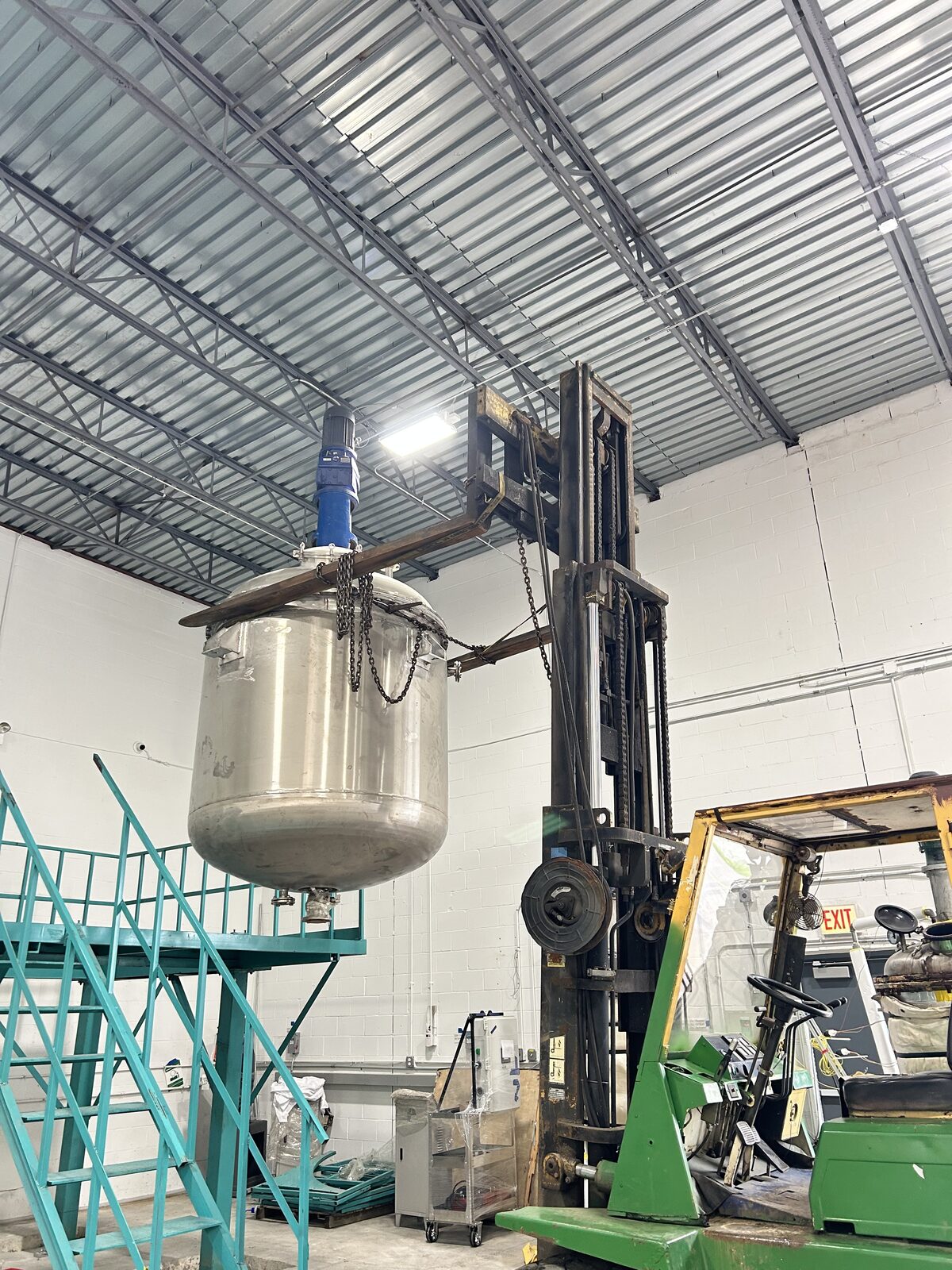 A forklift lifting a stainless steel mixing vessel on chains, beside a teal steel stairway