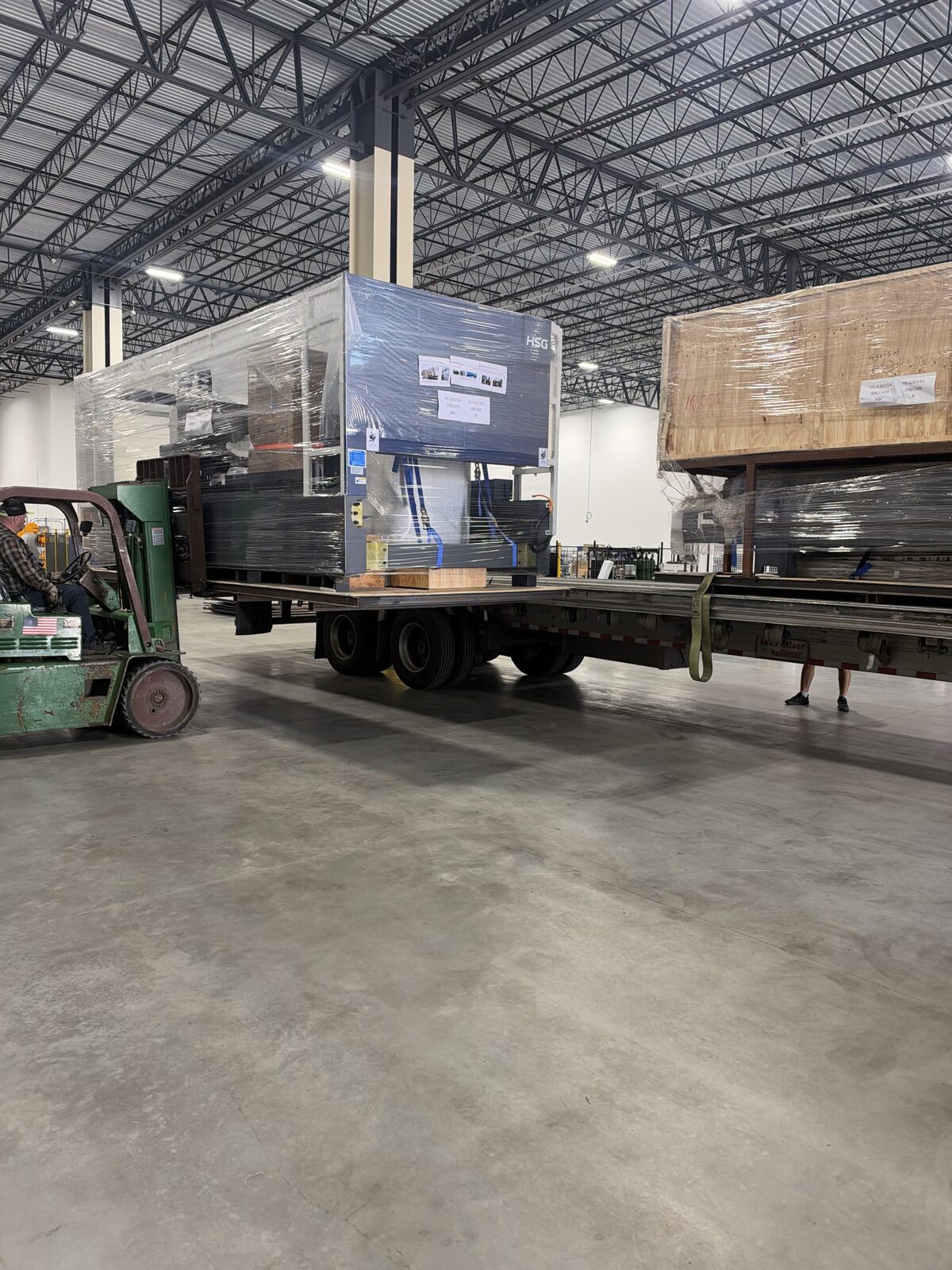 A forklift at the back of a trailer carrying a stretch-wrapped HSG laser machine, inside a warehouse