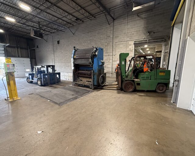 A green forklift carrying a press unit toward the dock, with our blue forklift at the left