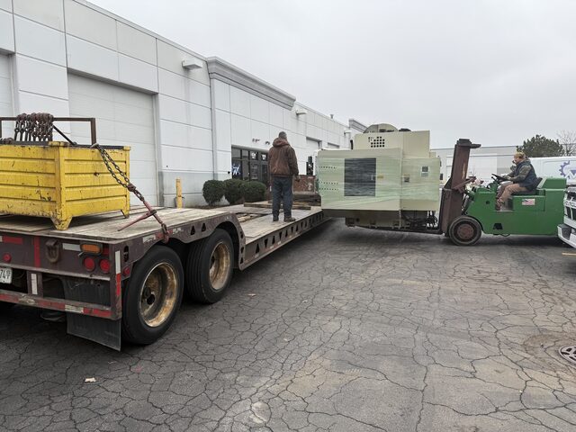 Green forklift carrying a wrapped CNC machine toward the back of a lowboy trailer