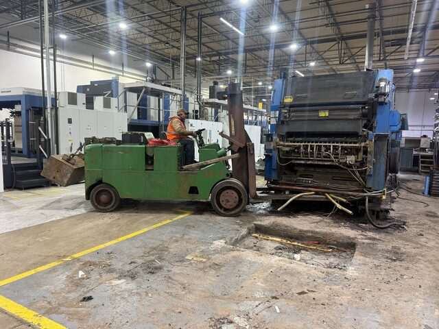 The operator on the forklift at the end press unit; a steel box hangs on a chain from the back of the forklift