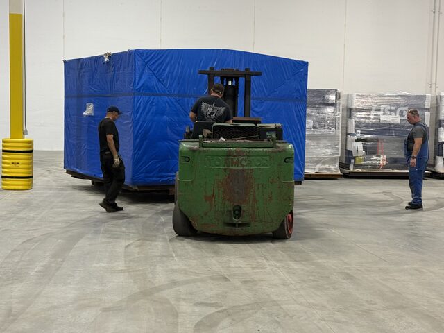 Our Towmotor forklift carrying a crate under a blue tarp, seen from behind; wrapped HSG crates stand by the wall