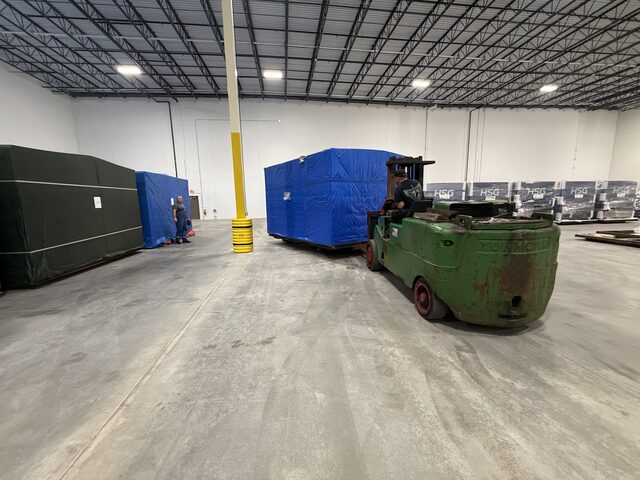 The forklift with the blue-tarped crate in a warehouse, with a row of HSG crates along the wall