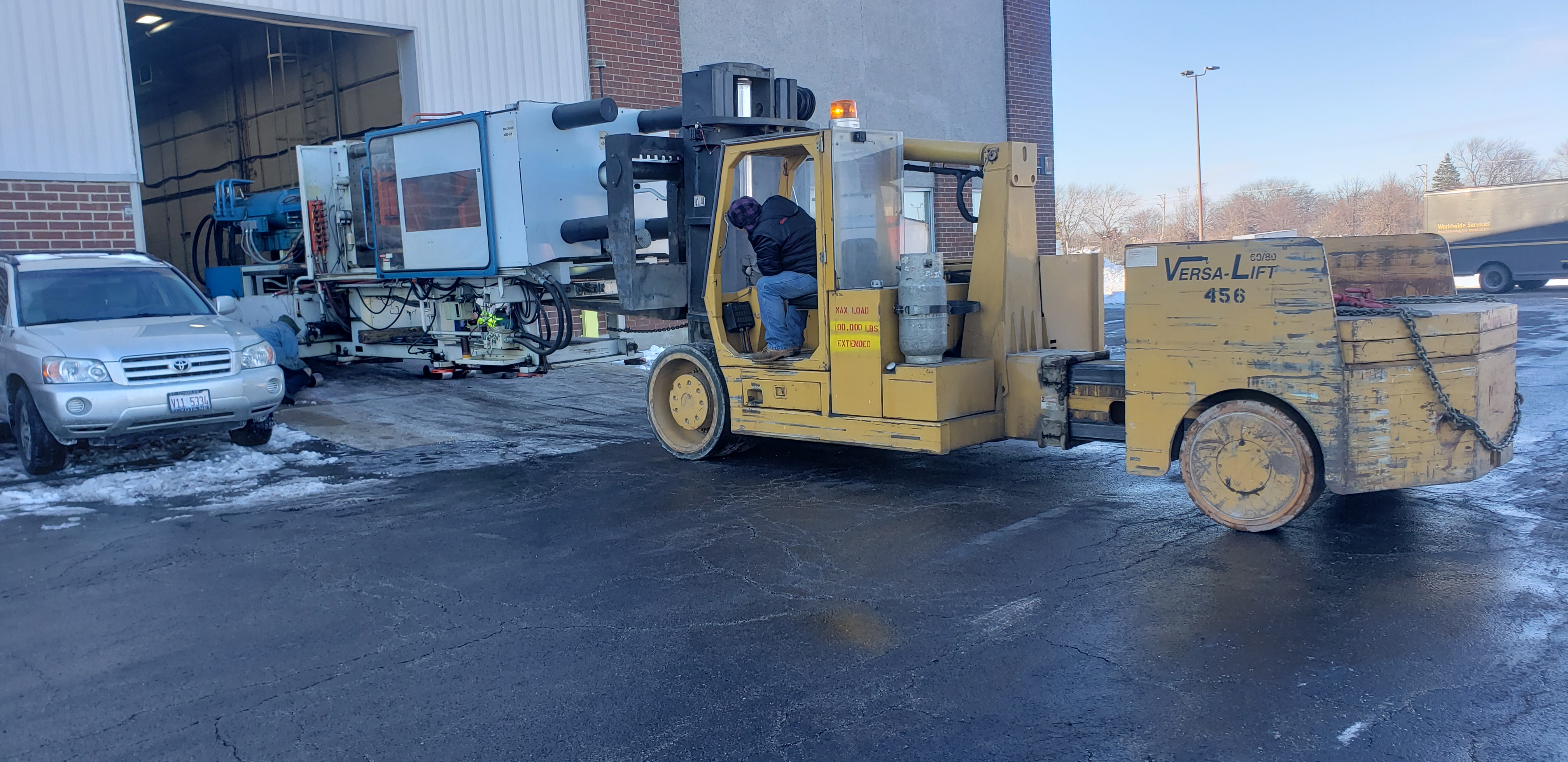 A yellow Versa-Lift 60/80 forklift with its forks under one end of a long injection molding machine, at an open overhead door, with snow on the ground